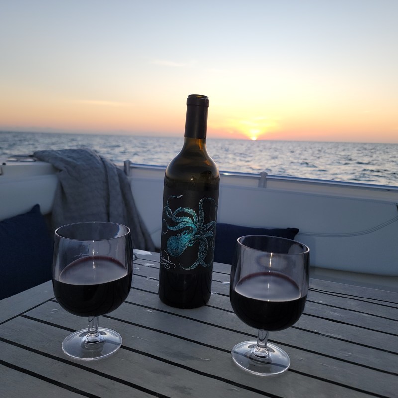 Wine bottle and two glasses on a boat deck at sunset over the ocean.